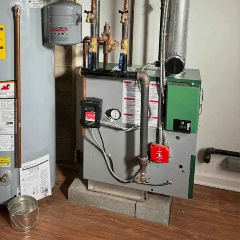 Residential gas-fired boiler with green metal housing, copper supply and return pipes, pressure gauge, and connected expansion tank. The boiler is installed on concrete blocks beside a water heater, with insulated vent pipes and control valves visible above.